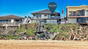 A coastal hotel with a modern design, featuring a prominent staircase leading to the entrance, a flag, and a clear view of the beach.