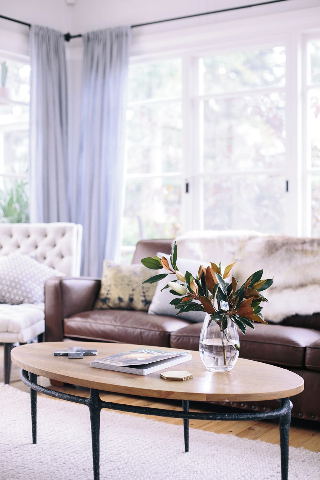 A living room with a round coffee table, a brown leather sofa, a tufted armchair, and a vase with green leaves.