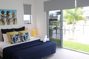 A bedroom with a large bed, a painting on the wall, and a view of the outside. - The Lotus - Elite Holiday Homes (Gold Coast)