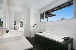 A modern bathroom with a large bathtub, two sinks, and a window providing a view of the sky. - Room for everyone in this luxurious waterfront home! (Gold Coast)