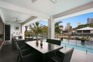A modern outdoor dining area with a pool, wicker furniture, and a city view. - Room for everyone in this luxurious waterfront home! (Gold Coast)