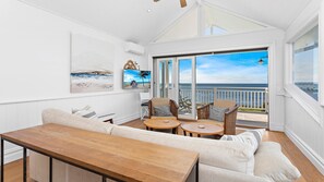 A spacious living area with a large sofa, two round wooden tables, and a wooden bench. There are two wicker chairs with cushions. The room has a large sliding glass door leading to a balcony with a view of the ocean. - Hamptons Escape - Magnificent views (Gerroa)
