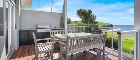A balcony with a wooden table and chairs, a grill, and a view of the ocean.
