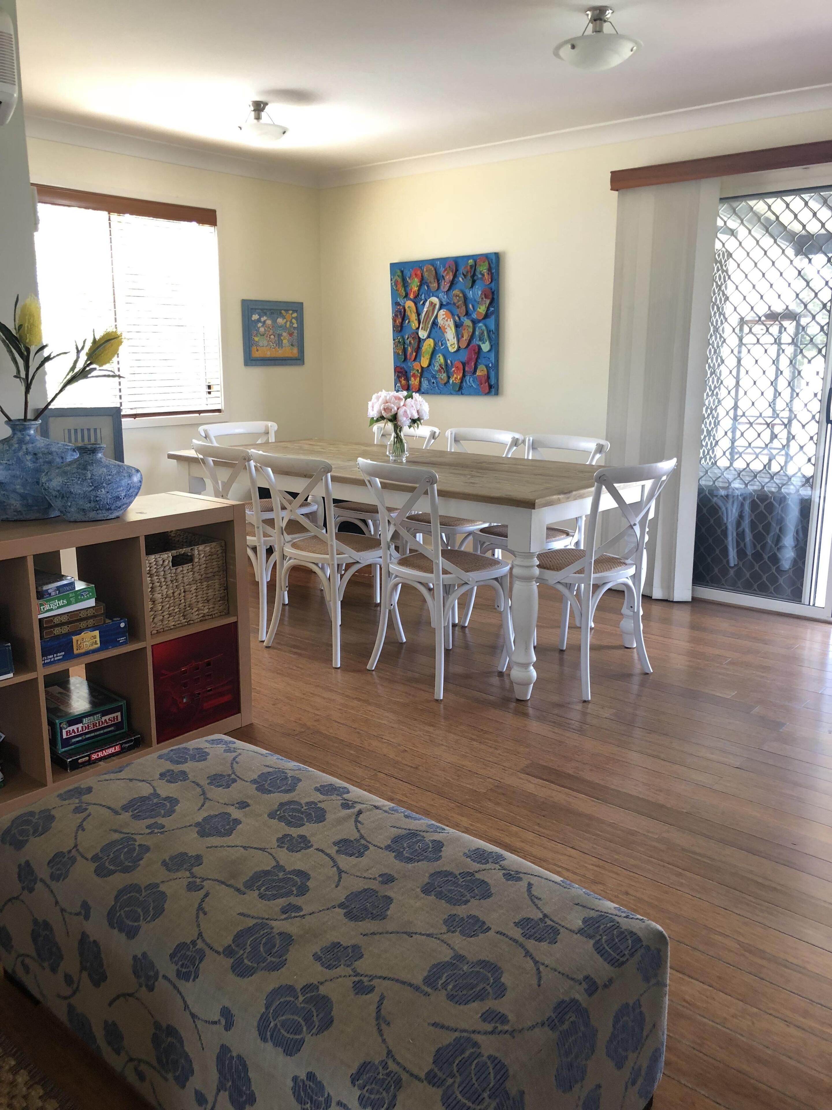 A dining area with a wooden table and white chairs, a vase of flowers, and a shelf with board games.