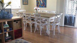 Een eetgedeelte met een houten tafel en witte stoelen, een bloemenvaas en een plank met gezelschapsspellen.
