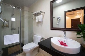 A bathroom with a glass shower enclosure, a toilet, a sink with rose petals, and a mirror. - Hanoi Lavender Hotel (Hanoi)