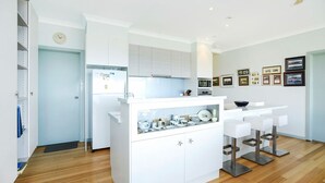 A modern kitchen with white cabinetry, a central island with bar stools, and a wall-mounted clock. - Magnificent Views Over the Bay (Encounter Bay)
