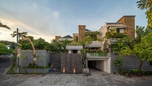 Een modern woongebouw met een gevel van natuursteen, een overdekte entree en een aangelegde tuin met tropische planten.