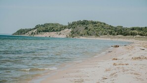 A beach with clear water, sandy shore, and a hill with trees. - Amazing Views All Seasons White Lake/Channel Spacious Easy Walk to Lake Michigan (Montague)
