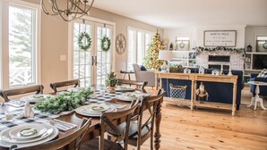 A dining area with a wooden table set for four, surrounded by chairs. A Christmas tree is visible in the background, and a fireplace is seen in the adjacent room. - Amazing Views All Seasons White Lake/Channel Spacious Easy Walk to Lake Michigan (Montague)