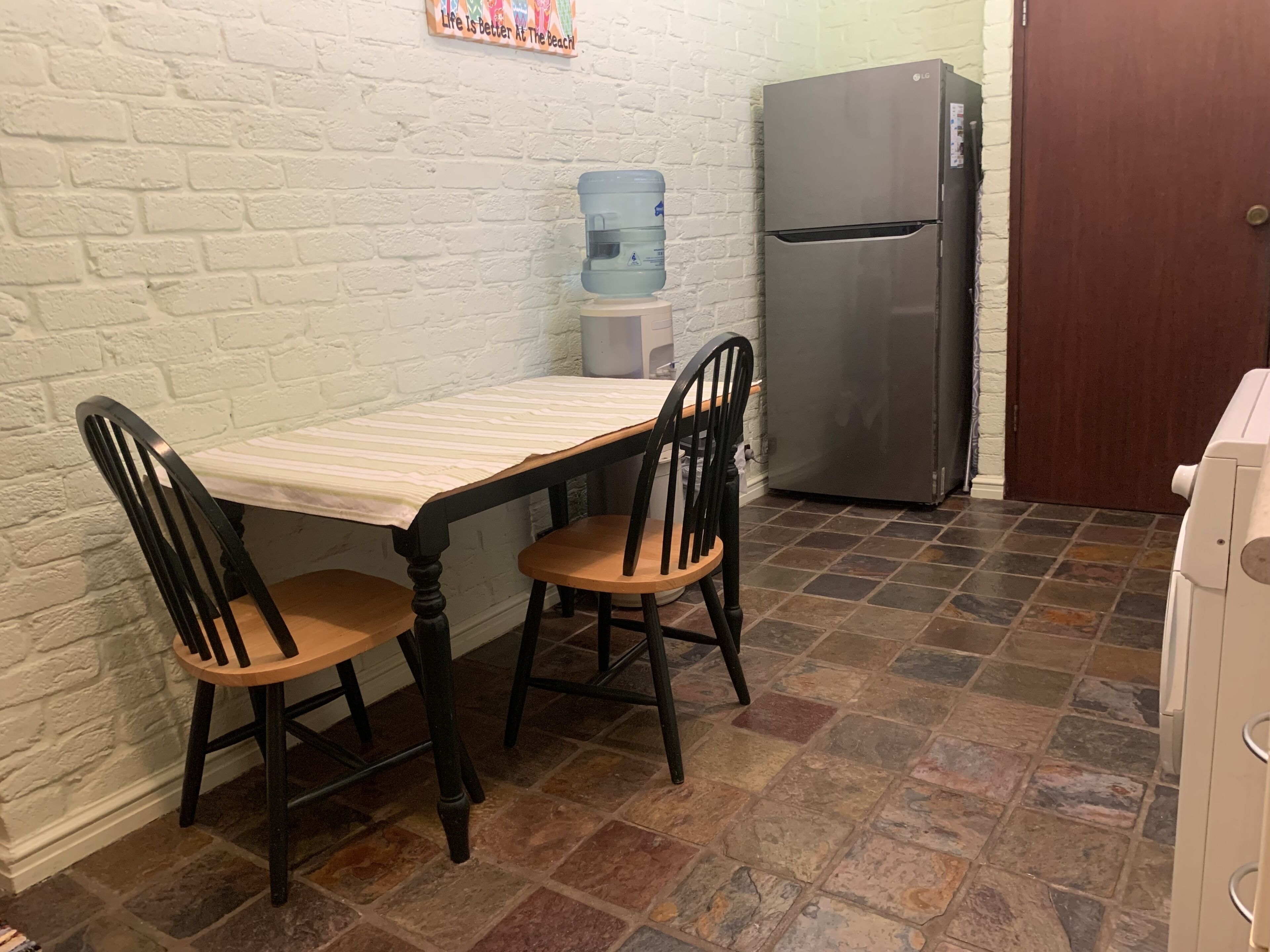 A kitchen with a white brick wall, a stainless steel refrigerator, a water dispenser, and a tiled floor.