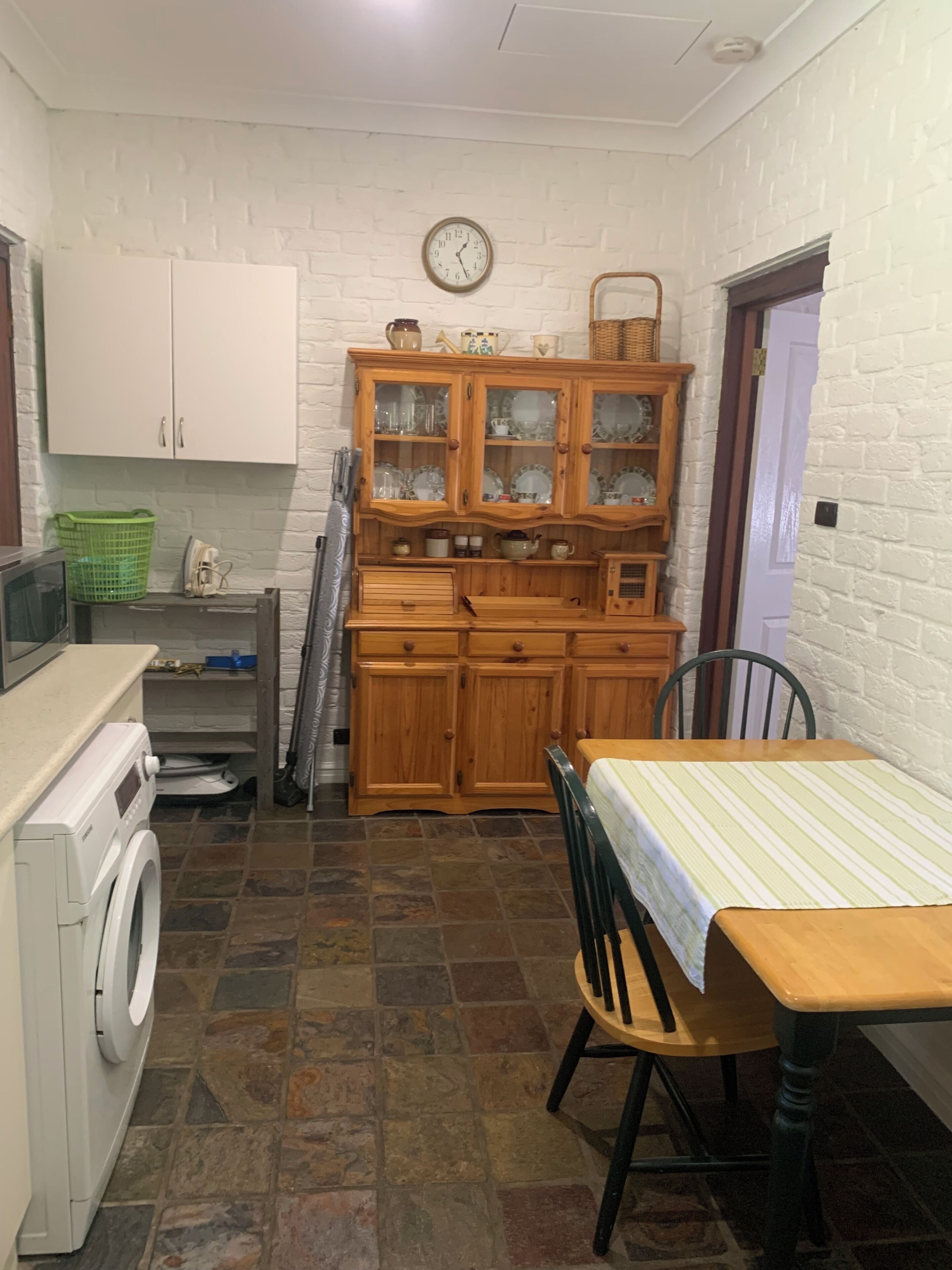 A compact kitchen with a washing machine, wooden cabinets, a dining table, and a clock on the wall.