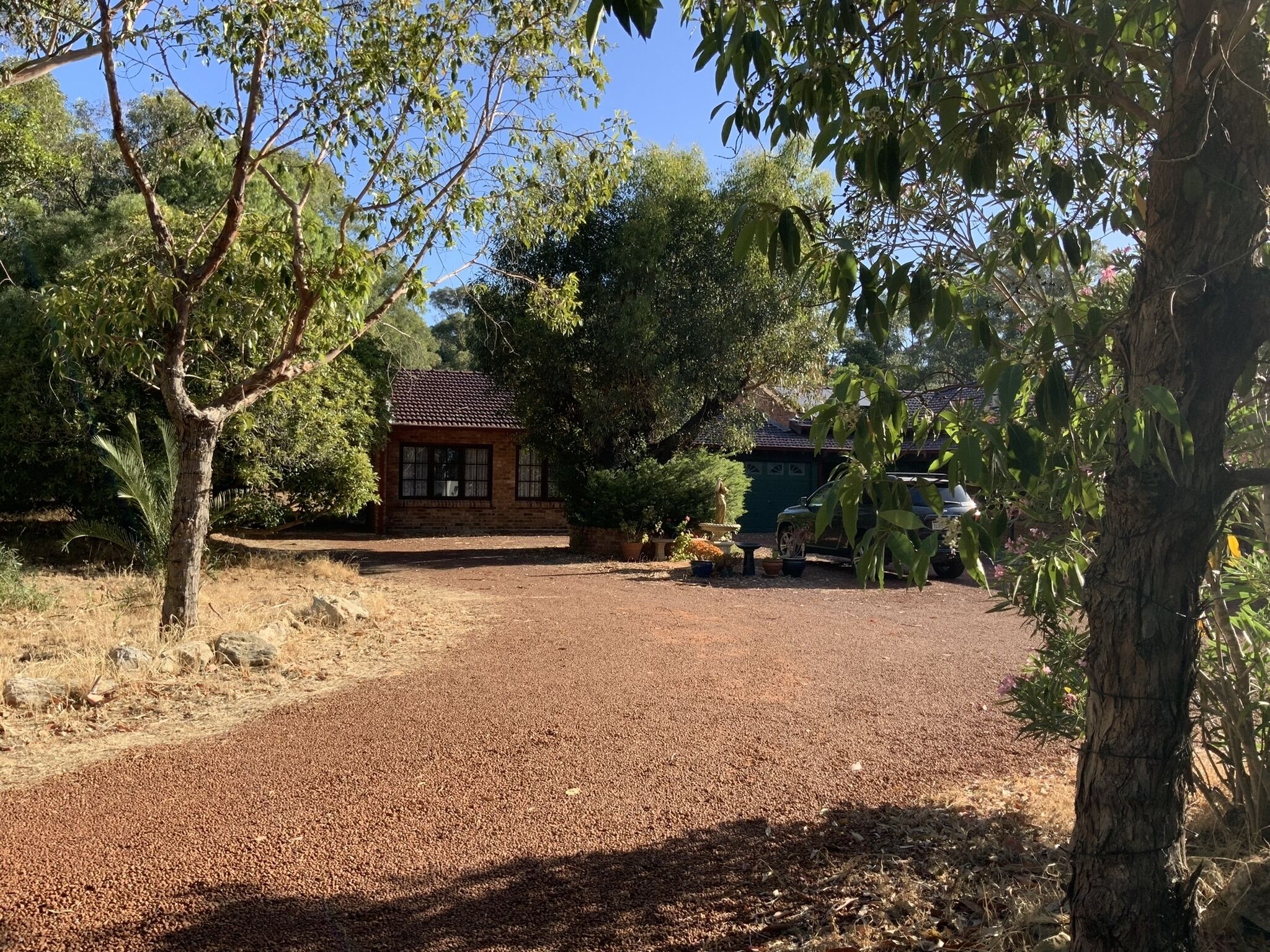 A gravel driveway leading to a brick house with a tiled roof, surrounded by trees and a garden.