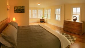 Ein Schlafzimmer mit Bett, Nachttischlampe, Kommode, Fenstern und Blick ins Freie.