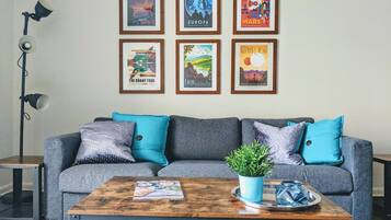 A living room with a grey sofa, blue cushions, a wooden coffee table with a plant, and framed posters on the wall.