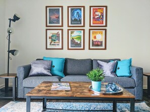 A living room with a grey sofa, blue cushions, a wooden coffee table with a plant, and framed posters on the wall. - Uptown Charlotte Pool View - 24/7 Fitness Center (Charlotte)