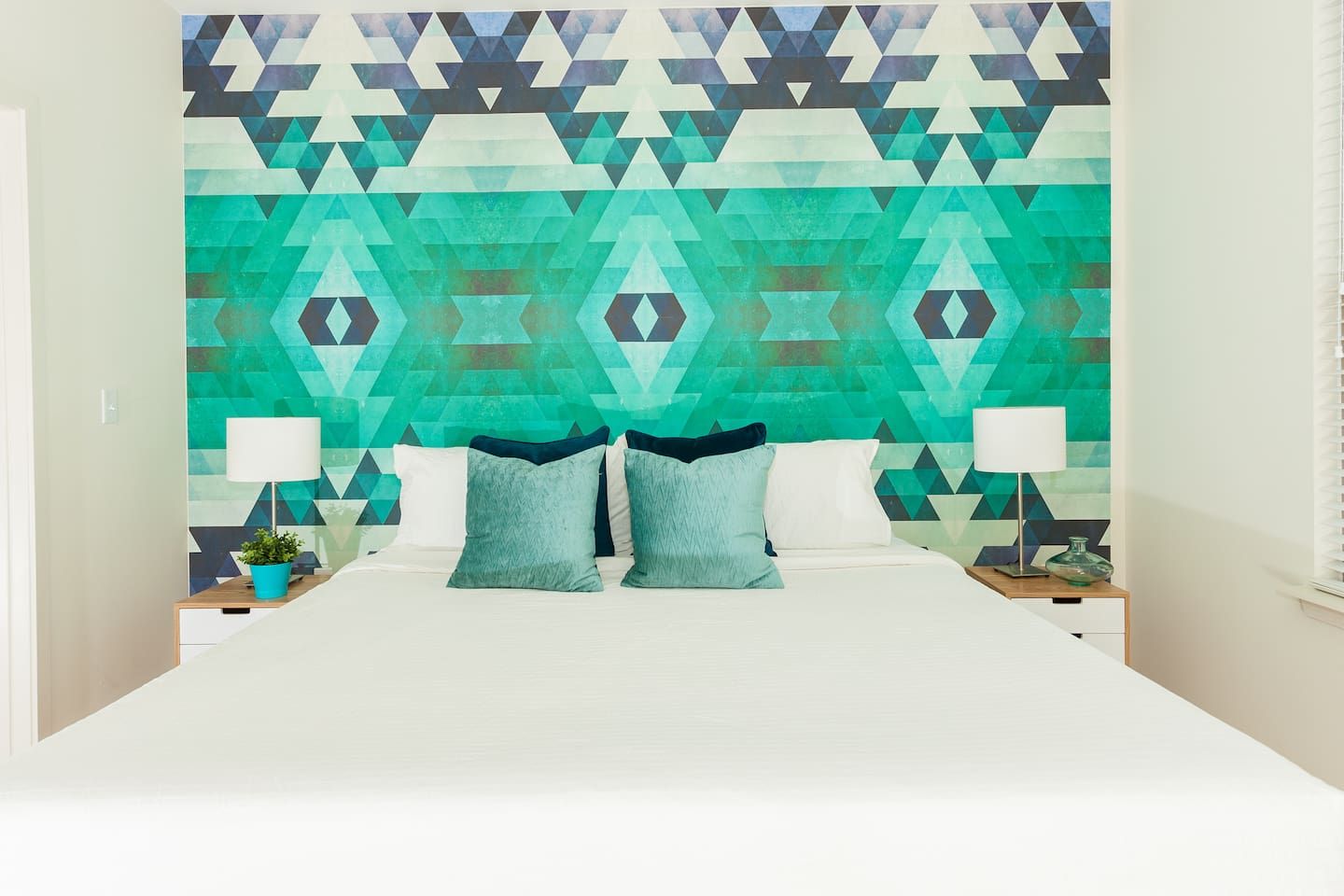 A bedroom with a geometric patterned wall, a bed with white bedding, two bedside tables with lamps, and a potted plant.