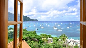 A view of a coastal area with numerous sailboats on the water, lush greenery, and a mountainous backdrop.