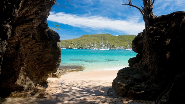 A beach with clear turquoise water, a sandy shore, and a view of boats and houses in the distance.