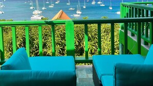 A balcony with two blue cushioned chairs, a green railing, and a view of the sea with sailboats.