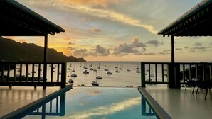 A poolside view with a sunset over the sea, boats, and a mountainous backdrop.