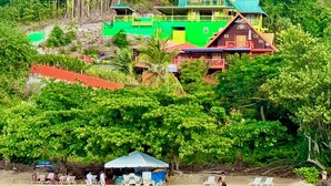 A large, multi-story house with a green roof and multiple balconies, surrounded by dense tropical vegetation.