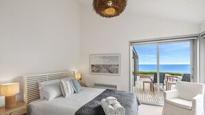 A bedroom with a bed, a nightstand, a lamp, a picture on the wall, and a view of the sea through a glass door. - The Cliff's Edge (Fairhaven)