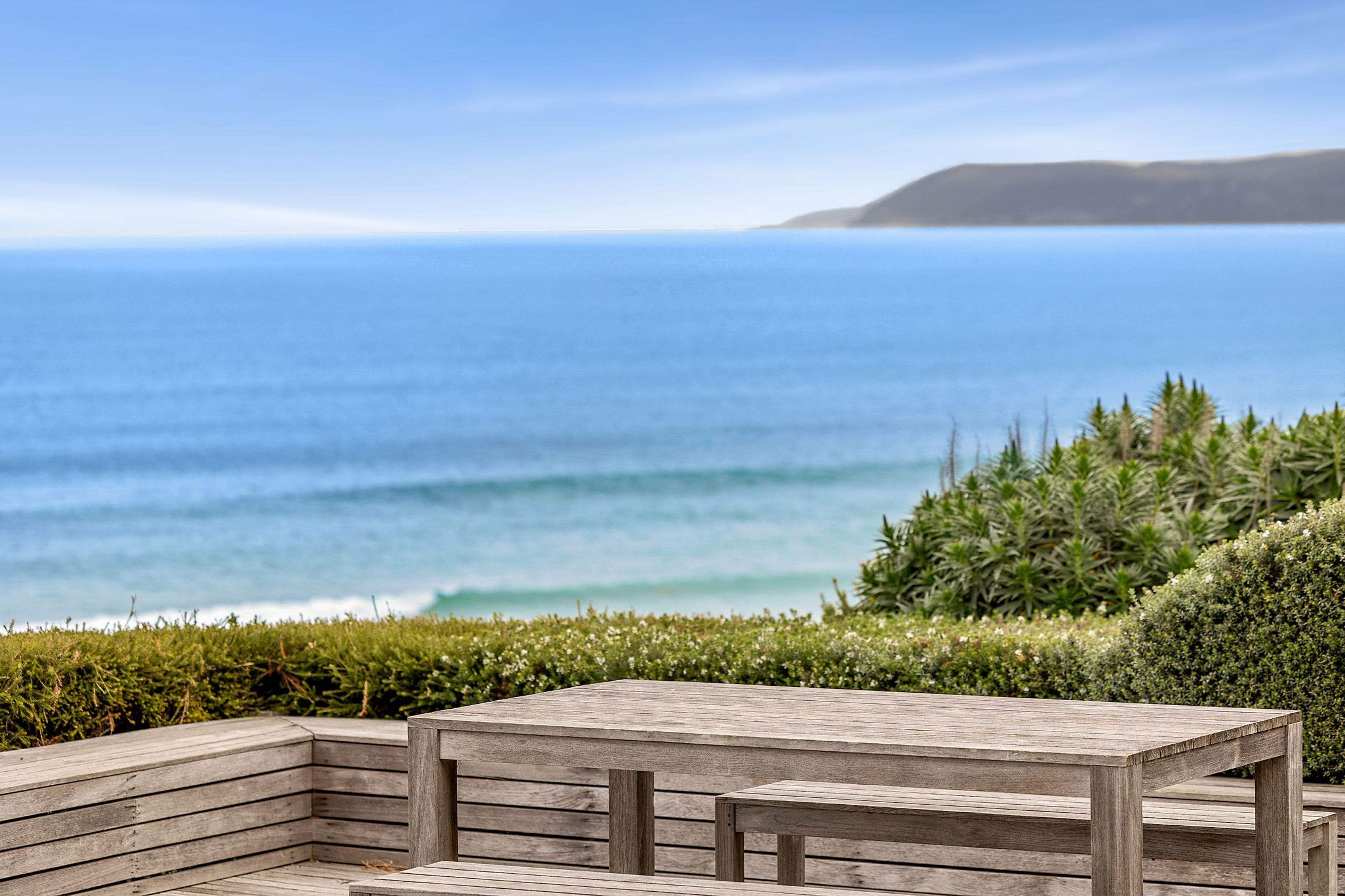 A wooden deck with a table and benches overlooking a beach with waves and a distant landmass.