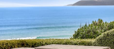 Une terrasse en bois avec une table et des bancs, donnant sur une plage où l’on peut voir des vagues et, au loin, une étendue de terre.