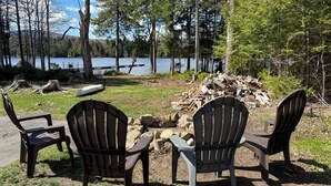 Un entorno junto al lago con dos sillas Adirondack mirando a un brasero, rodeado de árboles y una pila de leña.