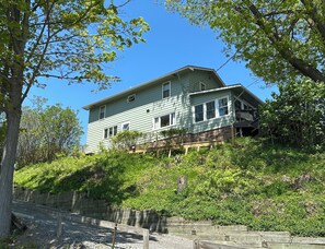 Une maison à deux étages, dotée d’un toit à forte pente, entourée d’une végétation luxuriante et d’arbres.