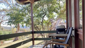 Eine Holzterrasse mit überdachter Sitzgelegenheit, einem Grill und Blick auf Bäume und Hügel.