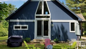 Een modern blauw huis met een brede voordeur, een klein portiek en een overdekte zitplaats met groene stoelen.