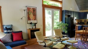 Een woonkamer met een blauwe bank, een bruin poef en een kamerkleed met een patroon. Er is een zithoek met een tafel en stoelen, en een keuken met een koelkast en kasten.