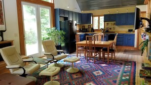 Een ruime woonkamer met een groot raam, een eethoek met een houten tafel en stoelen, en een keuken met blauwe kastjes en een zwarte koelkast.