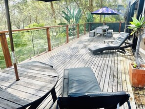 Um deck de madeira com cadeiras de descanso e uma mesa, cercado por guarda-corpos de vidro e vista para árvores.