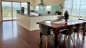 A spacious kitchen with a large island, a dining area with a wooden table and chairs, and a view through the sliding glass doors. - Large spacious house with many ocean views. Set in a peaceful quiet location. (Lake Bunga)