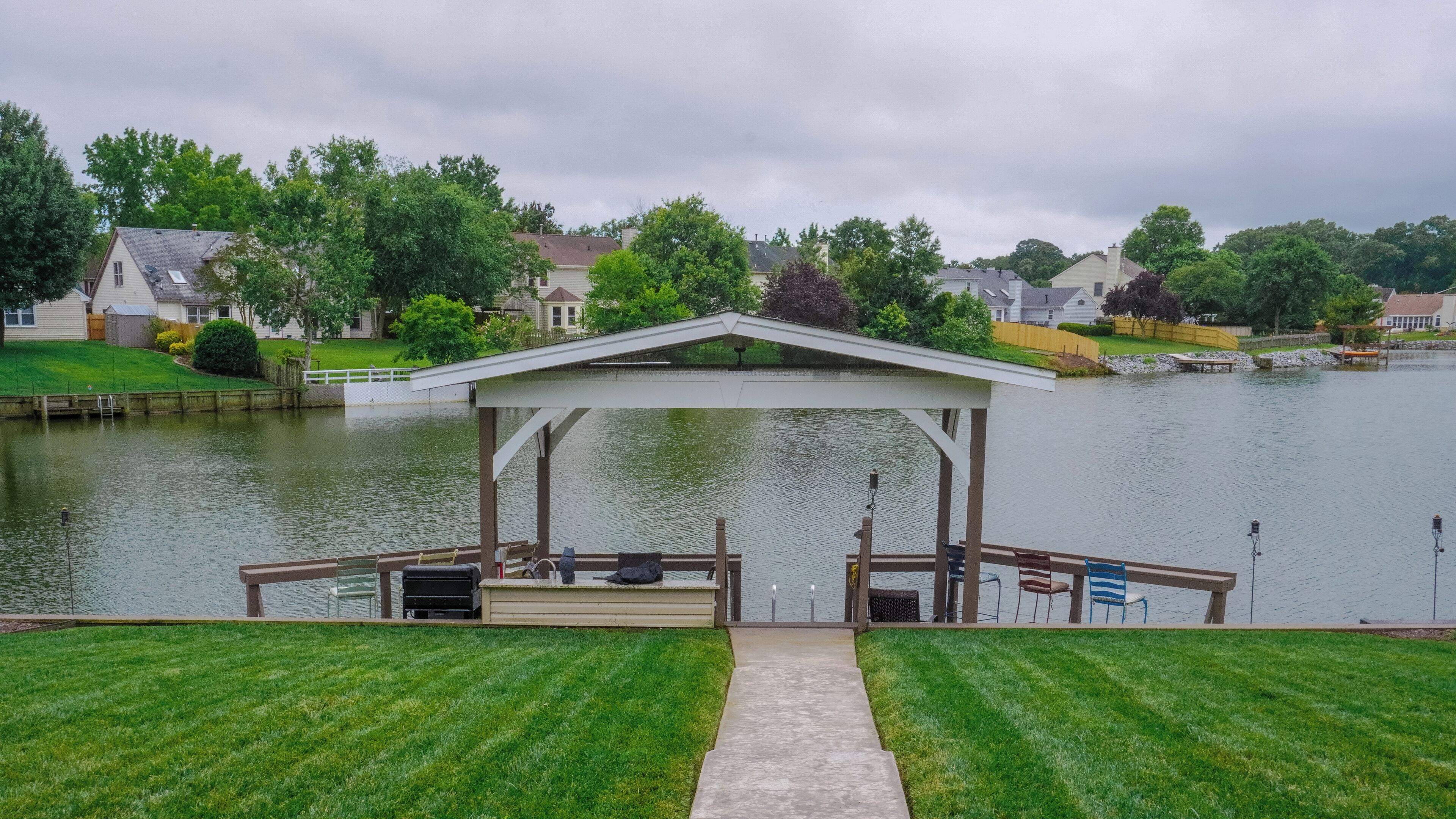 A gazebo with a dock and grill area overlooking a lake, surrounded by houses and greenery.