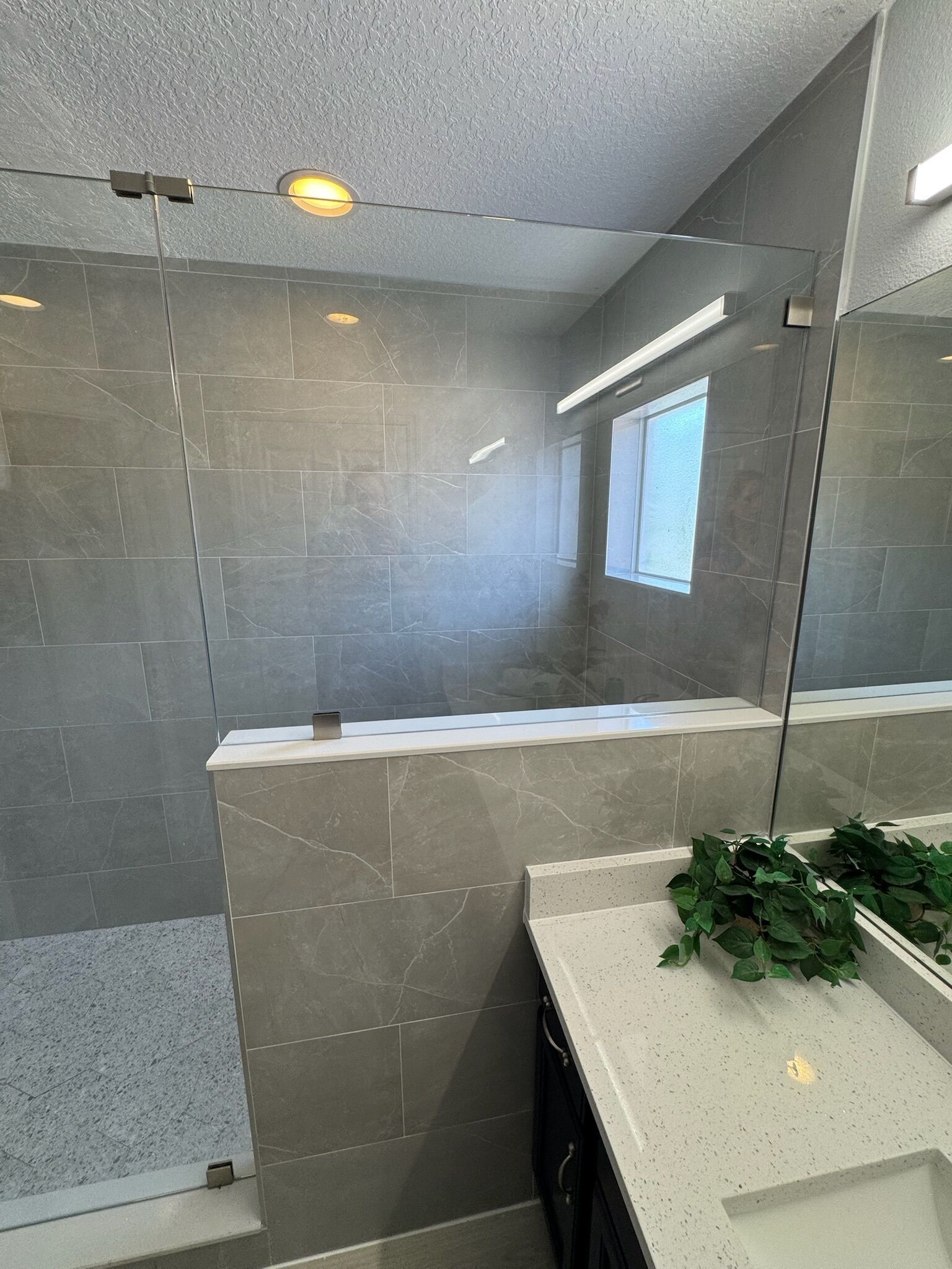 A modern bathroom with grey tiles, a glass shower enclosure, a window, and a plant on the counter.