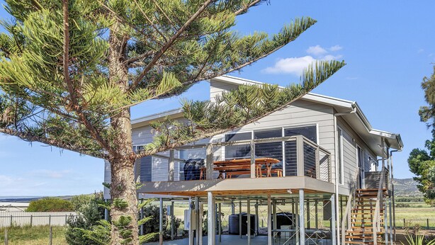 A modern two-story house with a covered patio, a wooden deck, and a large tree in the foreground. - Sea View at Silver Sands - Sitting Pretty with Sea, Hills and Washpool Views (Aldinga Beach)