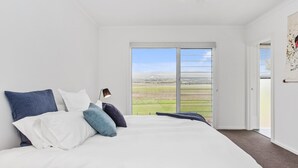 A bedroom with a large bed, a nightstand, a lamp, and a view of a green landscape through a sliding door. - Sea View at Silver Sands - Sitting Pretty with Sea, Hills and Washpool Views (Aldinga Beach)