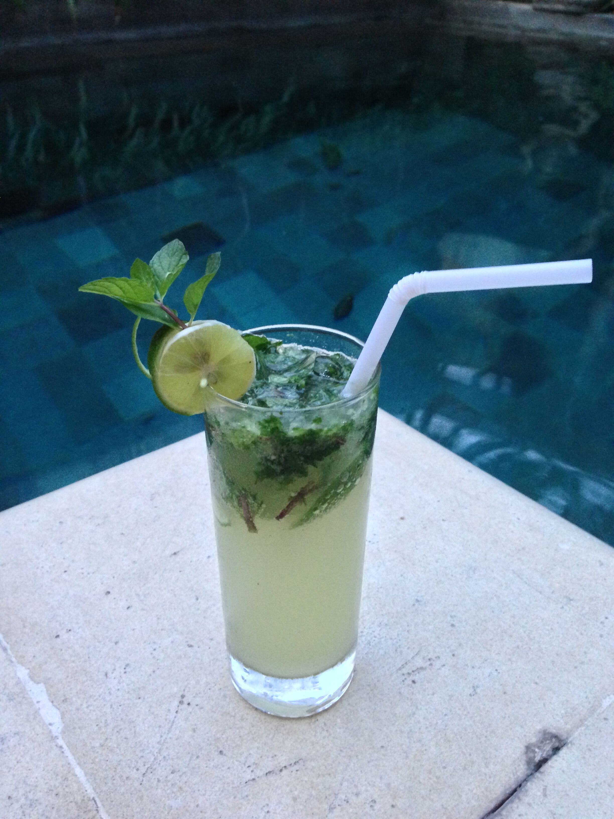 Un grand verre contenant une boisson claire, ornée d’une tranche de citron vert et de quelques feuilles de menthe, posé sur une surface en pierre, près d’une piscine.