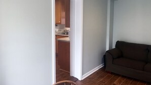 A living room with a dark brown sofa, a ceiling fan, and a kitchen visible through an open doorway.