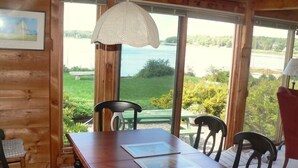 Una mesa de comedor de madera con sillas, un cojín rojo y vistas a un lago a través de amplios ventanales.