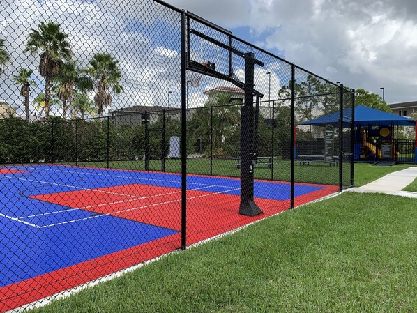 Una cancha de baloncesto rodeada de césped verde y palmeras, con una superficie de juego azul y roja.