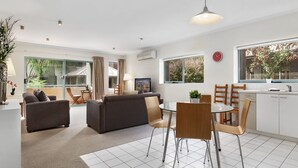 A modern living room with a sofa, a dining table with chairs, a kitchen area, and large windows with a view of greenery. - Cumberland Resort Lorne private Apartment near beach, pool, spa, tennis, shops (Lorne)