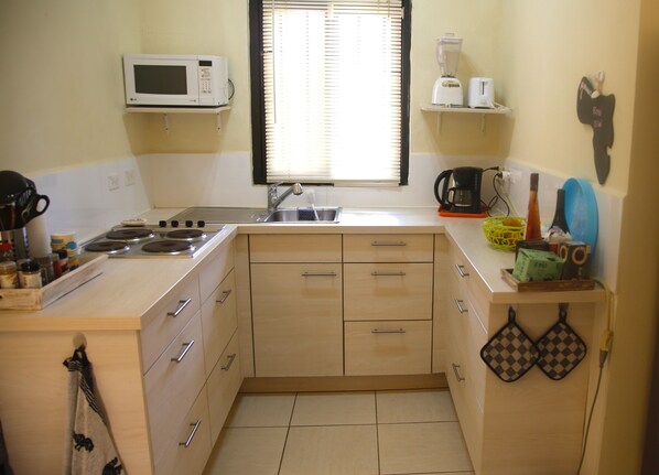 A compact kitchen with a microwave, stove, sink, and a window with blinds. - Your Windsurf Home Away From Home (Kralendijk)