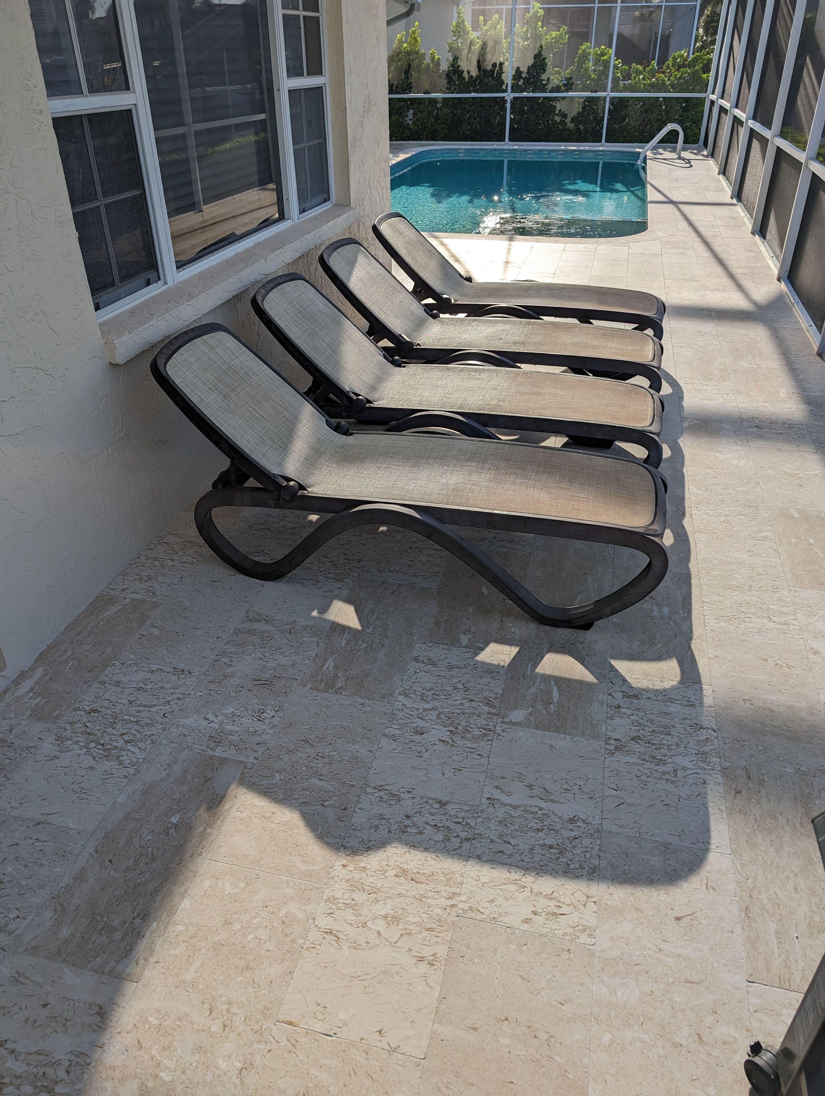 A poolside area with four sun loungers aligned against a wall, a pool in the background, and a view of greenery beyond.