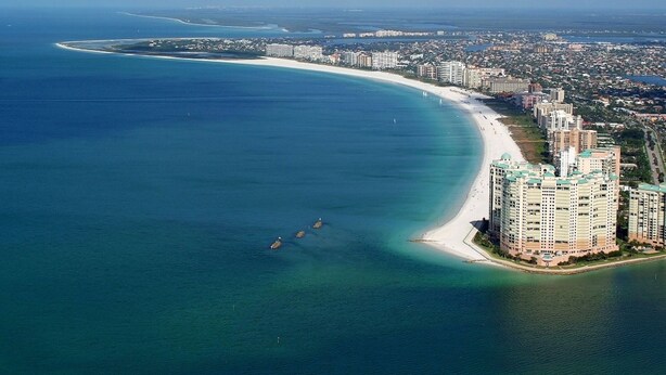An aerial view of a coastal city with a long sandy beach, a marina, and high-rise buildings along the shore. - Avail August 28 & on. Sunny New heated Pool & Spa 10min walk2beach 6-bikes (Marco Island)