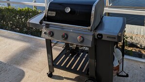 A portable Genesis II grill with four burners, a thermometer, and a side burner, set up on a patio with a view of a lake and residential buildings. - Avail August 28 & on. Sunny New heated Pool & Spa 10min walk2beach 6-bikes (Marco Island)